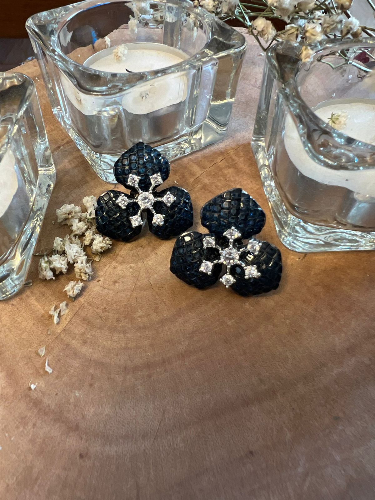 Black three-petal earrings with textured surface and central crystal floral motif, placed on wood with glass containers and dried flower fragments.