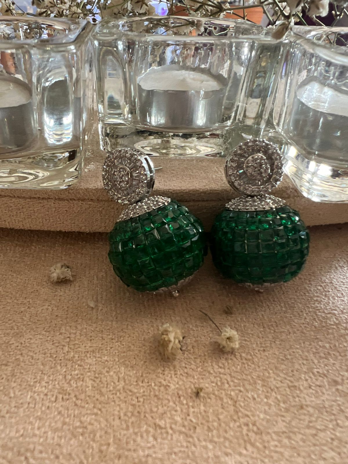 Ornate earrings with green square-cut stones in spherical design and halo of clear crystals, placed on beige surface with dried flowers and glass décor.