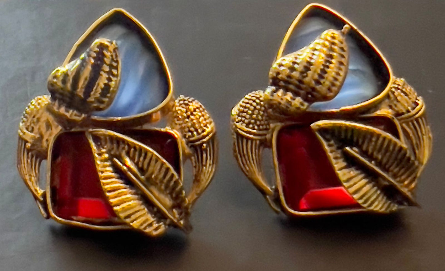 This image displays a pair of elegant earrings named "Amna." The earrings feature a striking combination of light blue and red semi-precious stones, intricately set in a copper-based metal with detailed gold accents.