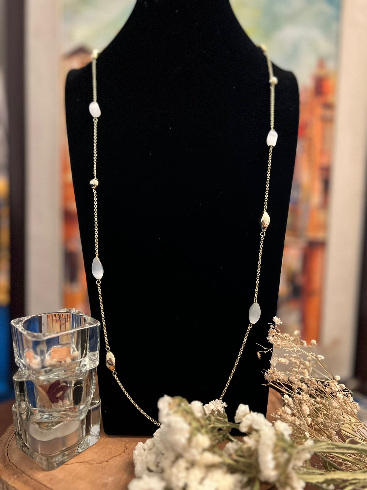A silver-colored necklace with white bead charms displayed on a mannequin bust, with decorative dry flowers and a glass object partially visible in the foreground.