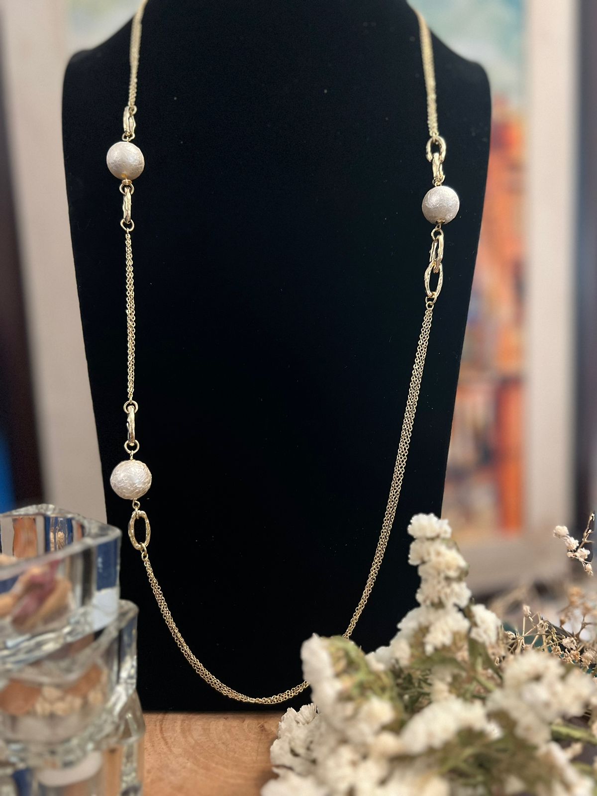 A gold-colored thick chain long necklace with white pearl-like beads displayed on a mannequin bust, with a floral arrangement in front.