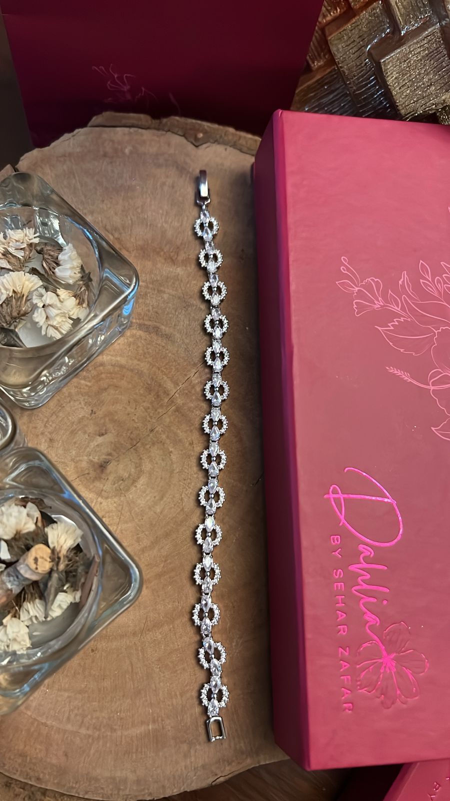 A bracelet with silver-colored beads placed on a wooden surface next to a red box and a glass vase with decorative items.
