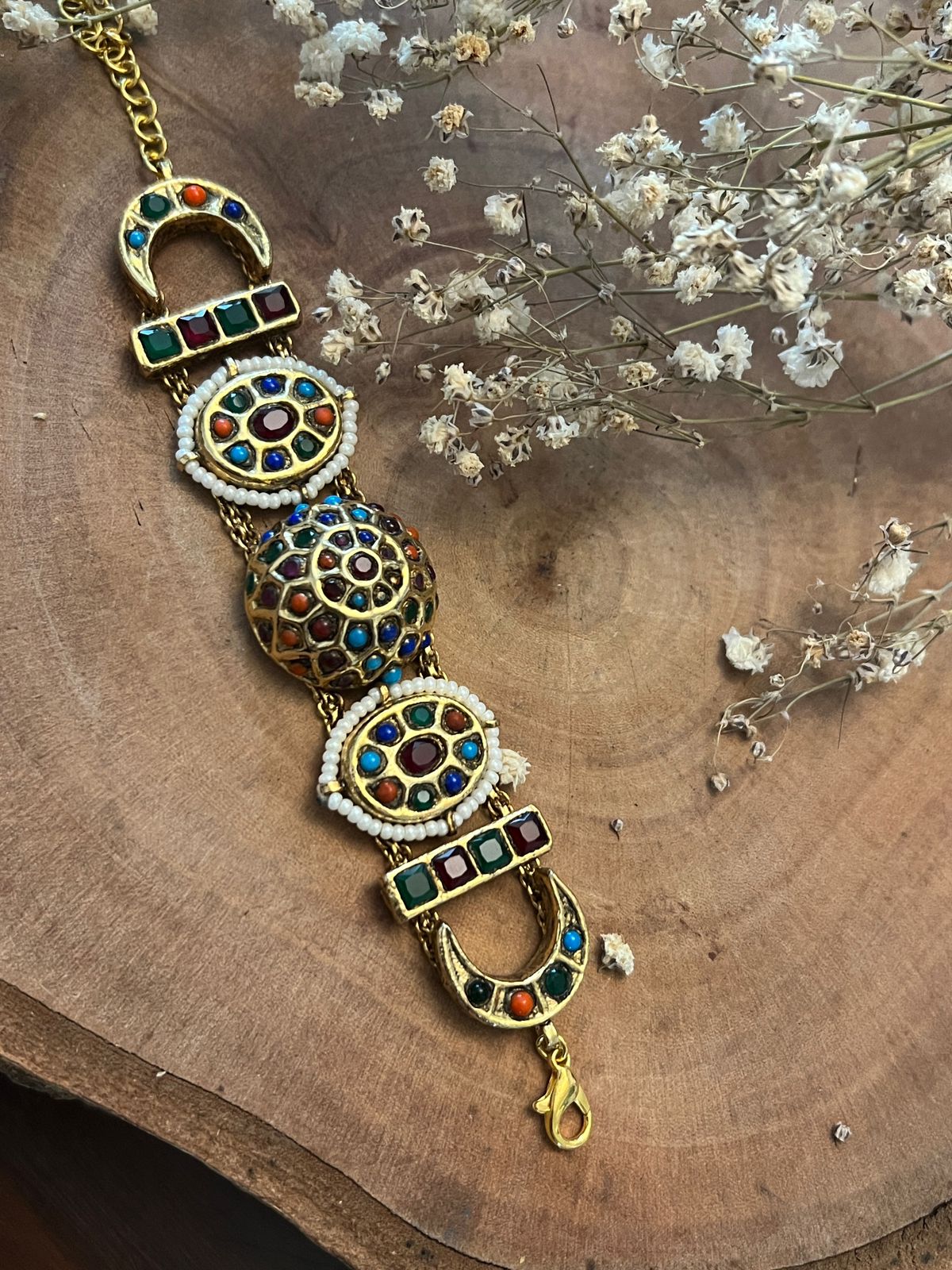 A multicolored floral patterned bracelet with gold accents and intricate detailing, displayed on a wooden surface with a background of dried flowers.