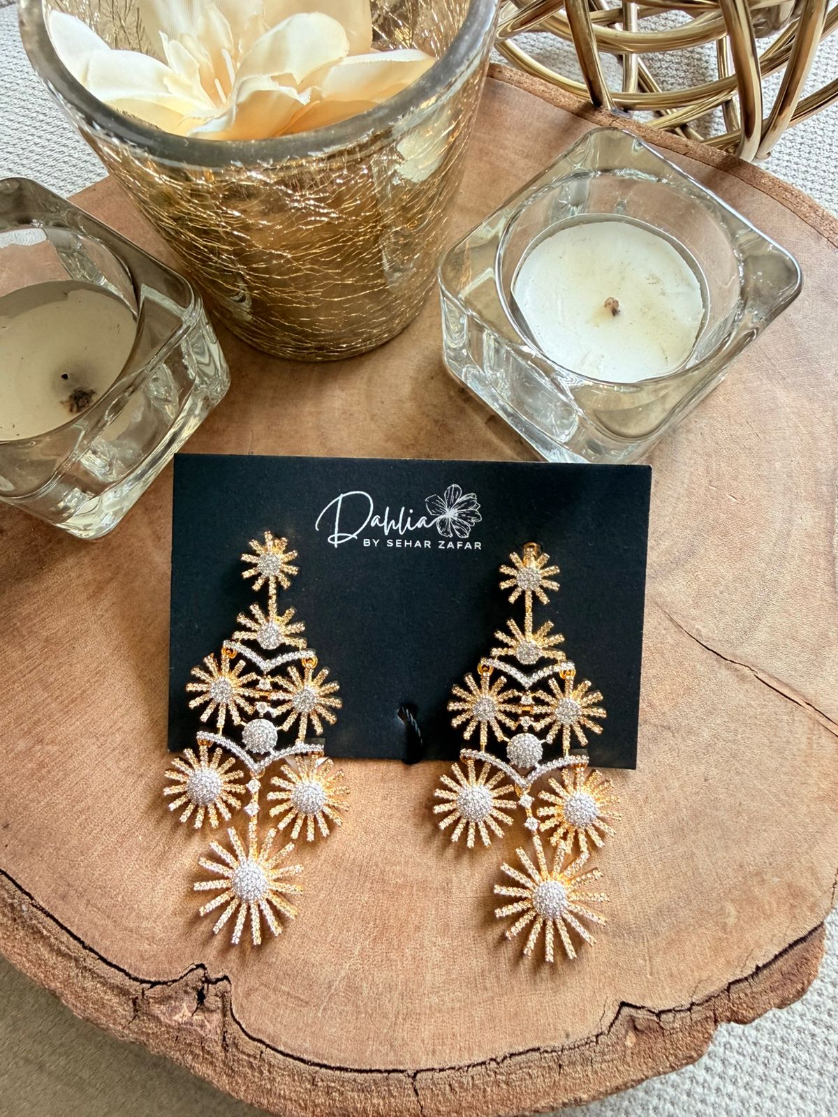 Gold and white earrings on a black card with 'Deshani' branding, placed on a wooden surface with candles and a vase.