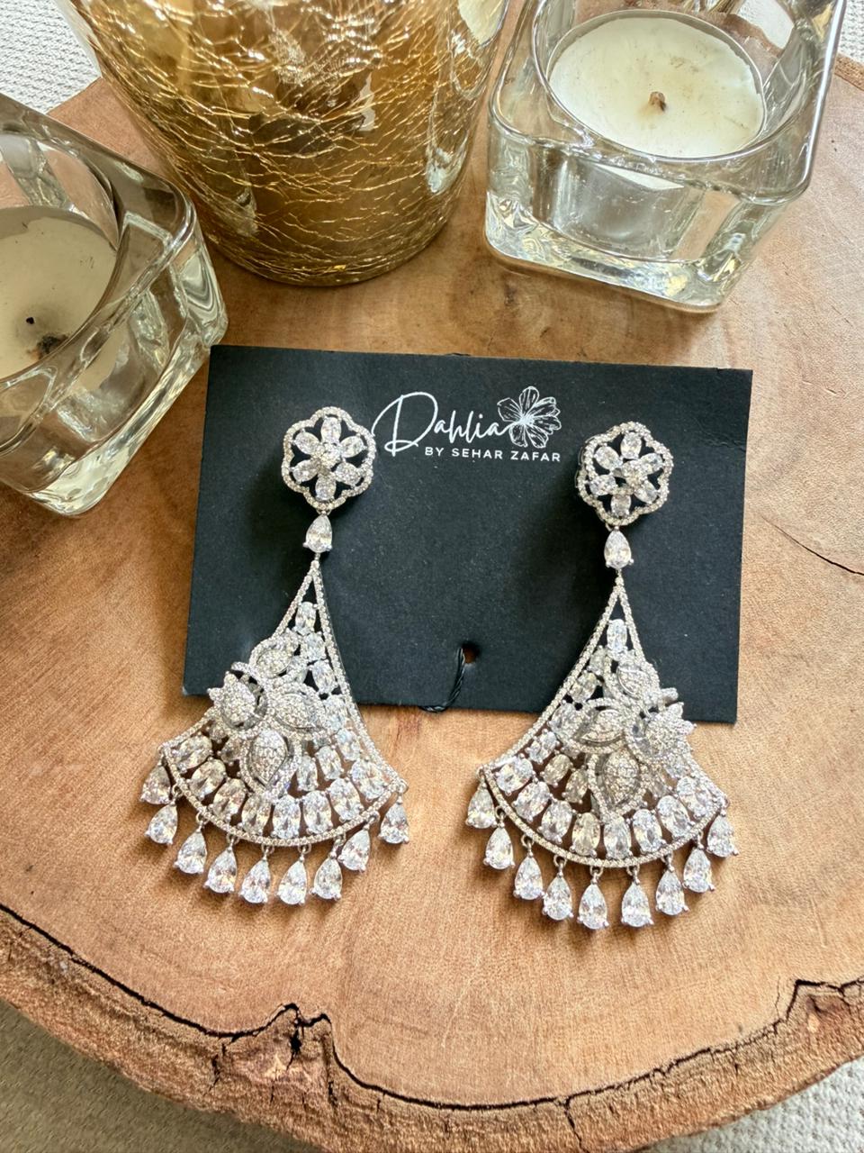 Decorative earrings on a black card with 'Dhara' branding, placed on a wooden surface with candles.