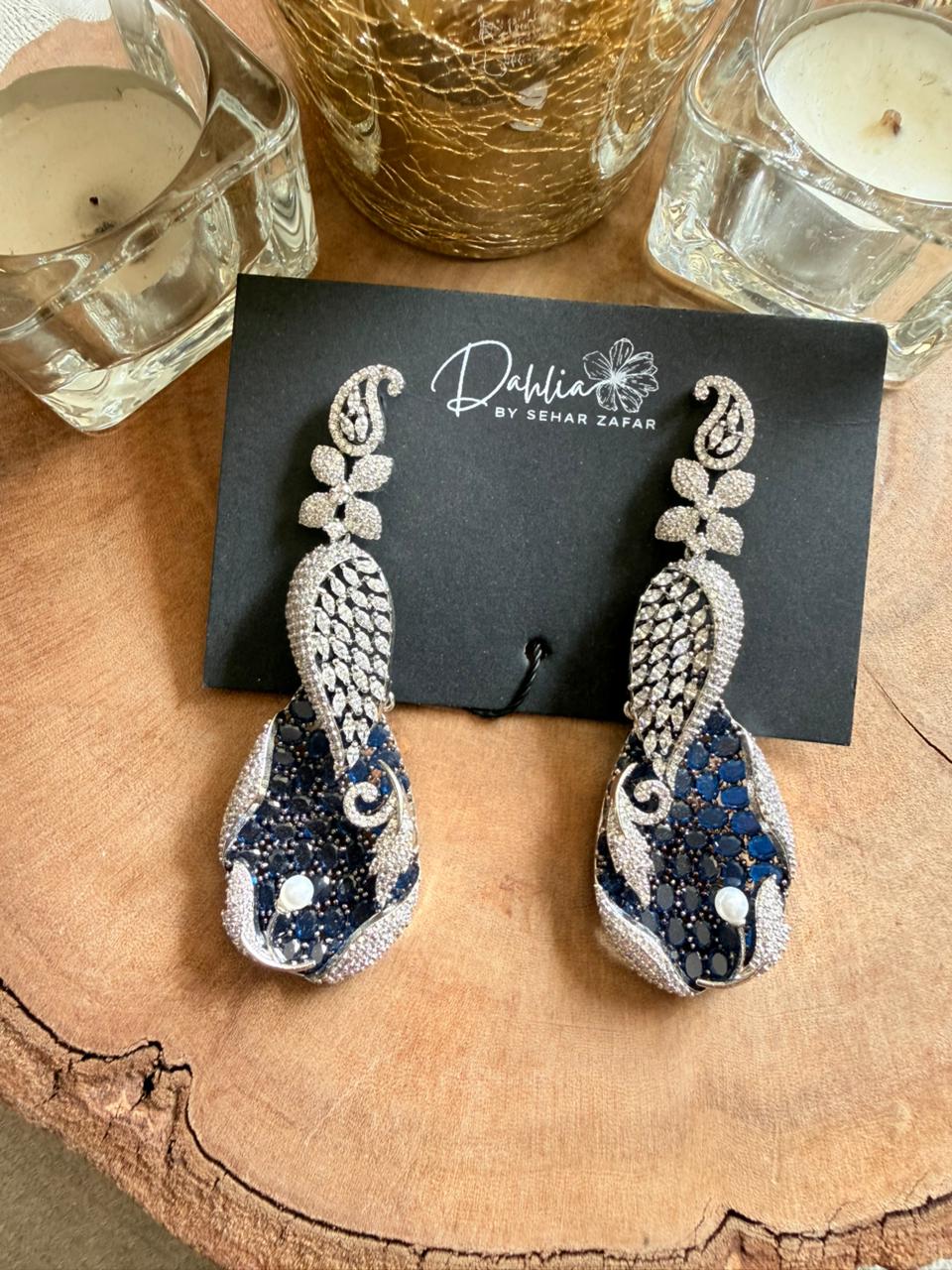 Decorative earrings on a black card with 'Dolce' branding, placed on a wooden surface with candles in the background.