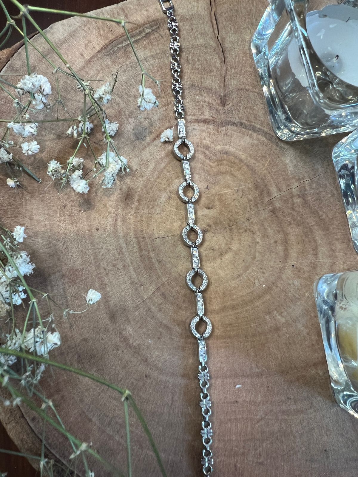 Silver bracelet with circular and rectangular links, some adorned with stones, placed on rustic wood with white flowers and glass candle holders.