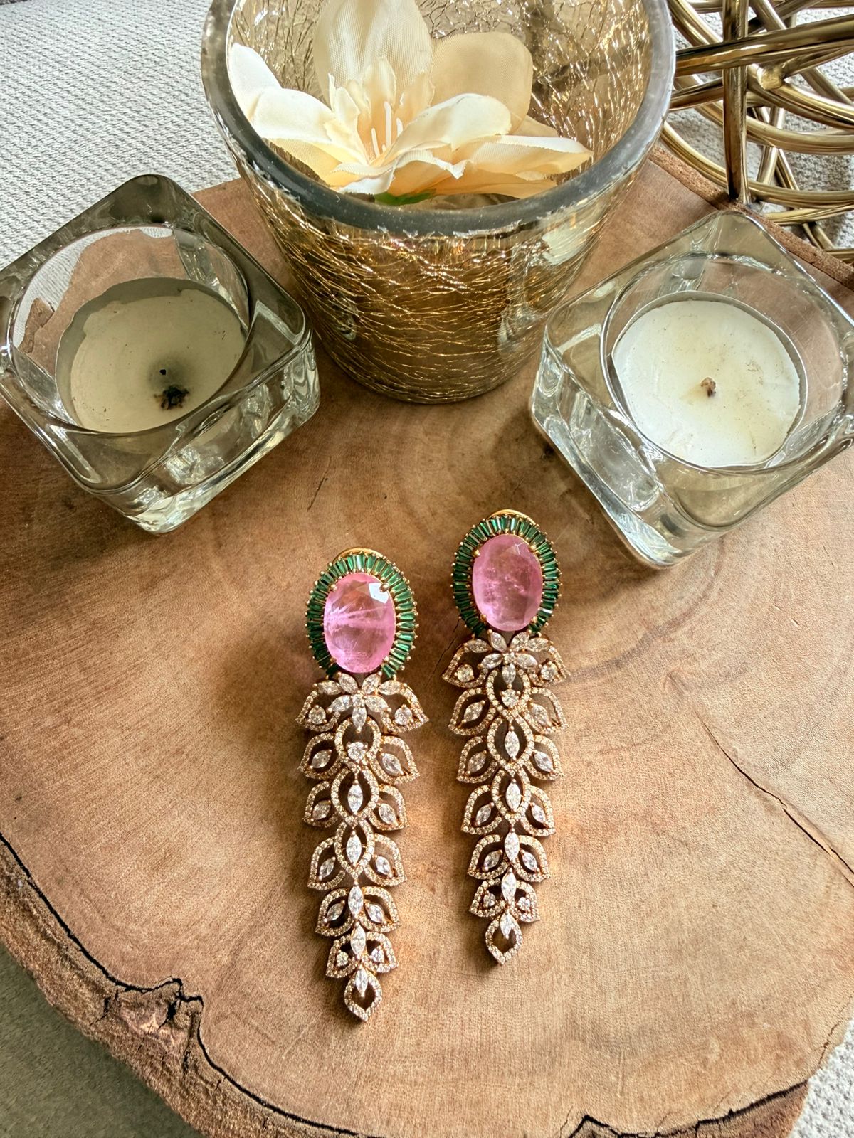 Ornate earrings with pink gemstones on a wooden surface with candles and flowers.