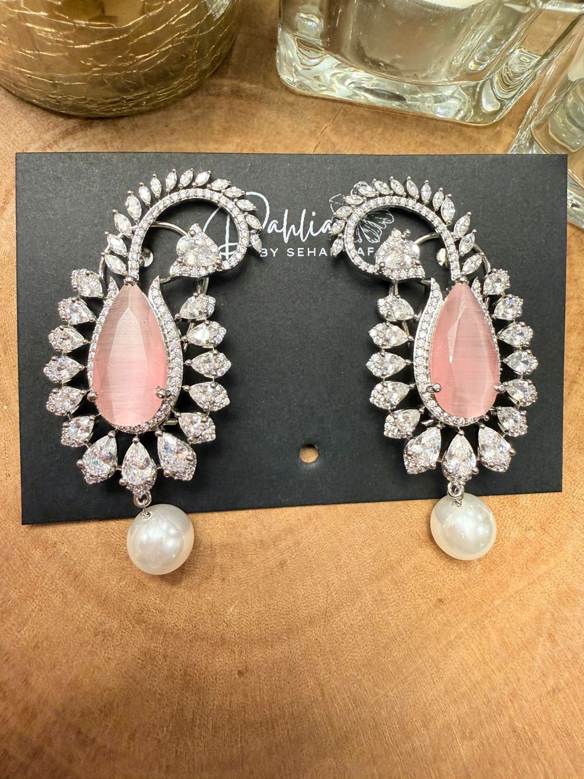 Pair of earrings with pink stones and pearl drops on a black card with 'Sahil by Sehaj' branding, placed on a wooden surface.
