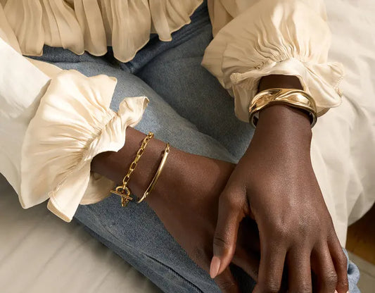 Close-up of hands resting on denim jeans, styled with gold metallic bracelets and a neutral long-sleeve top, photographed