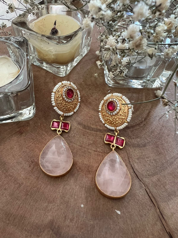 Ornate earrings with red and pink gemstones, gold circular tops with white beads, and teardrop pink stones, placed on wood with glass candle holders and dried flowers.