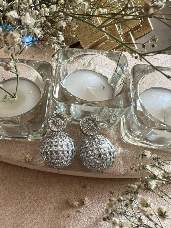 This image features a pair of elegant silver earrings, the Azra model, displayed alongside a serene setting of lit candles and delicate baby's breath flowers. The earrings are designed with a copper-inspired base, giving them a warm, lustrous finish.