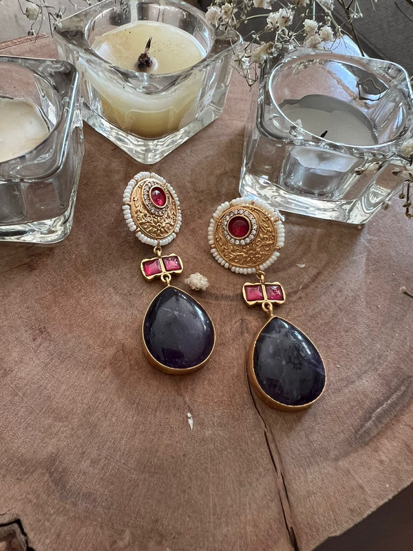 This image features a pair of exquisite earrings named "Mina," showcasing a blend of traditional craftsmanship and modern elegance. The earrings are crafted with gold-plated bases and adorned with intricate Meena work, complemented by semi-precious stones in shades of purple and red.
