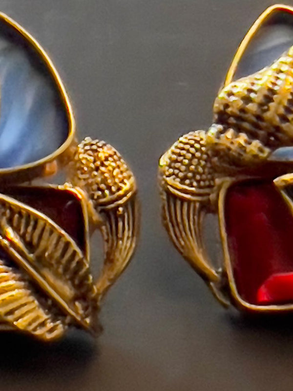 This image displays a pair of elegant earrings named "Amna." The earrings feature a striking combination of light blue and red semi-precious stones, intricately set in a copper-based metal with detailed gold accents.