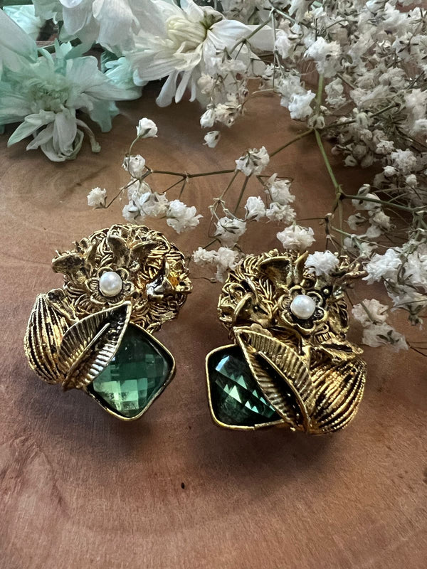 A pair of gold-colored earrings featuring green semi-precious stones and pearl details, displayed with white flowers.