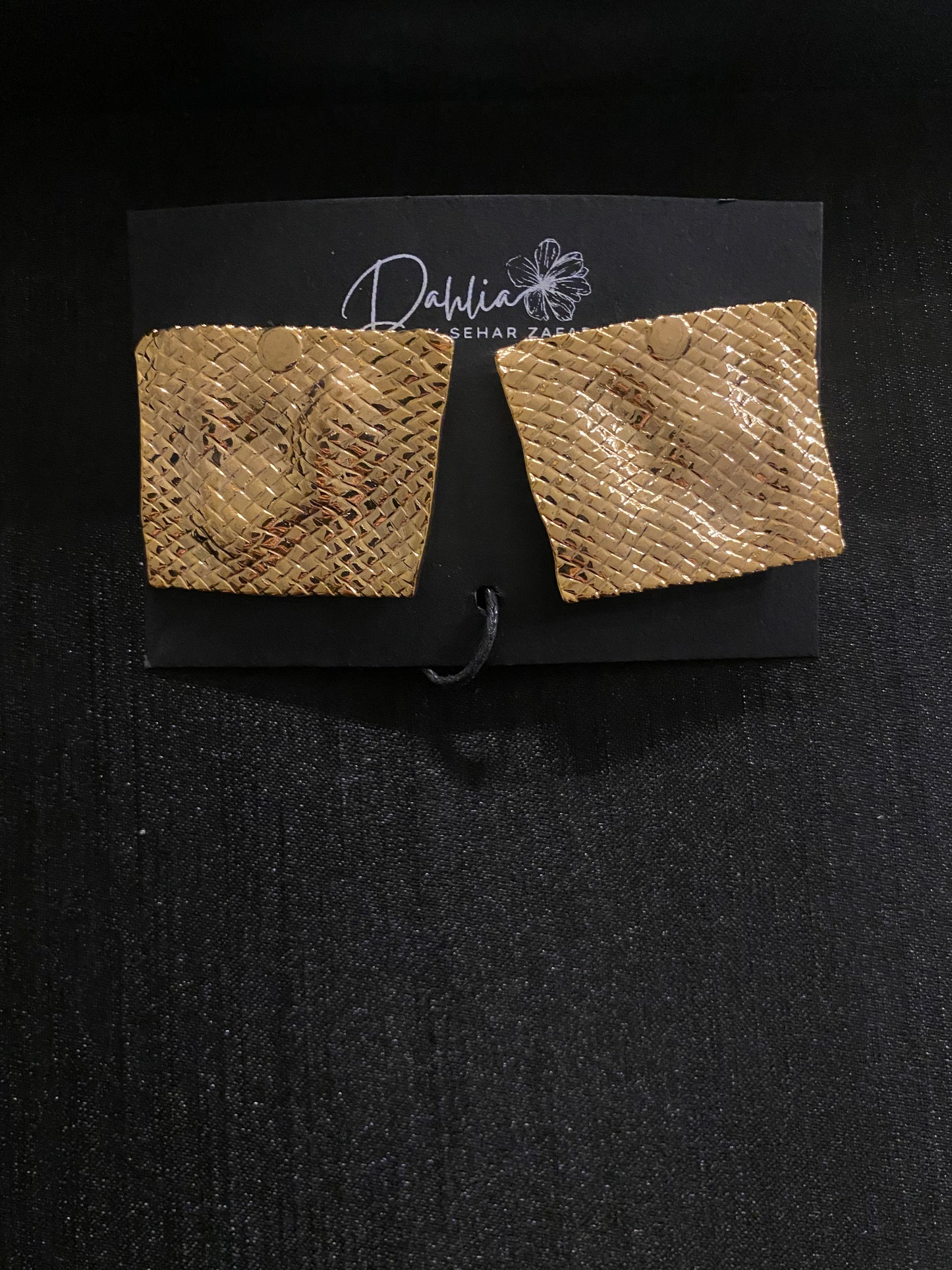 Gold square earrings with woven texture and curved shape on black branded card