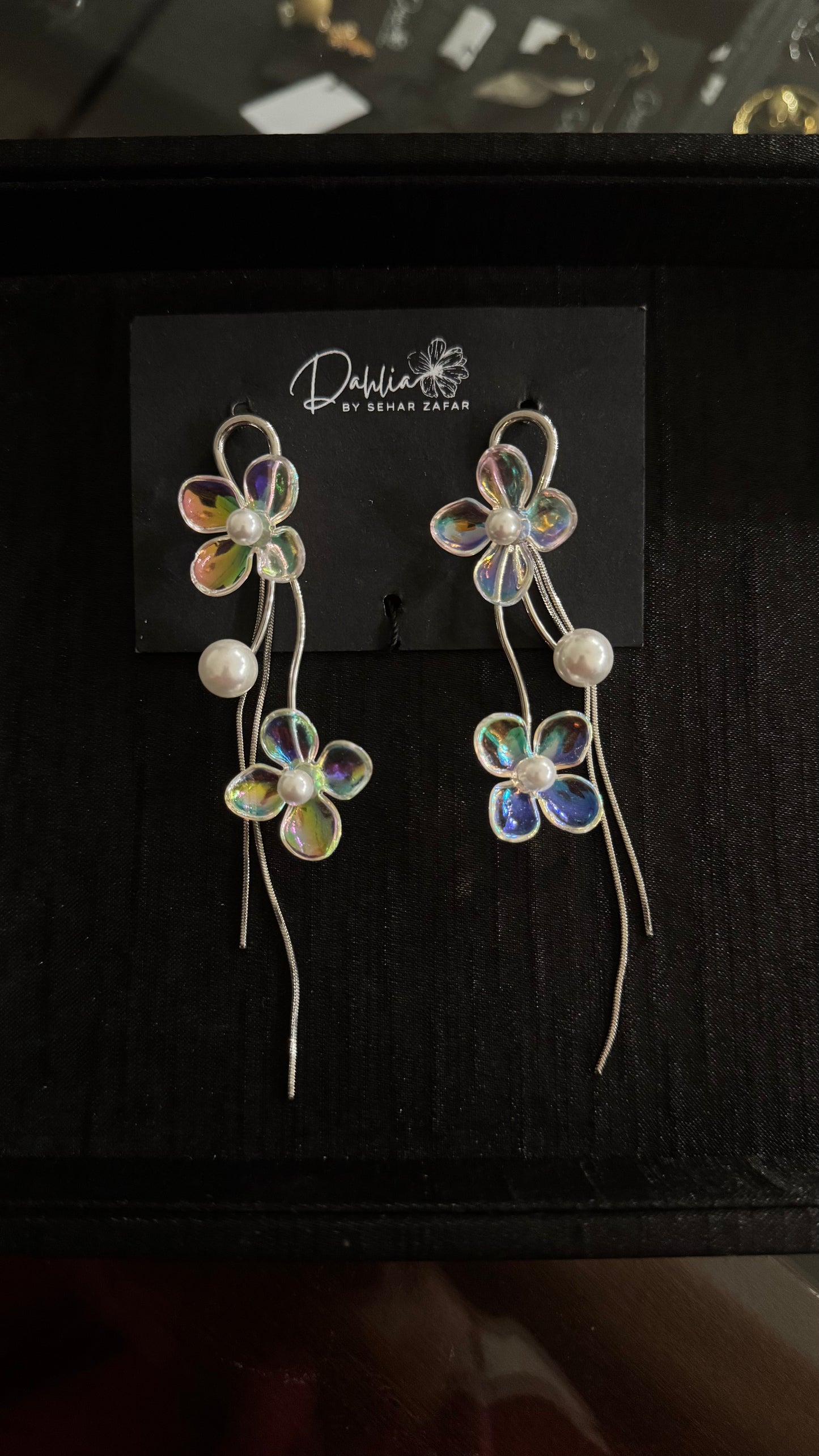 Iridescent floral earrings with pearl accents and curved metallic strands on black branded card
