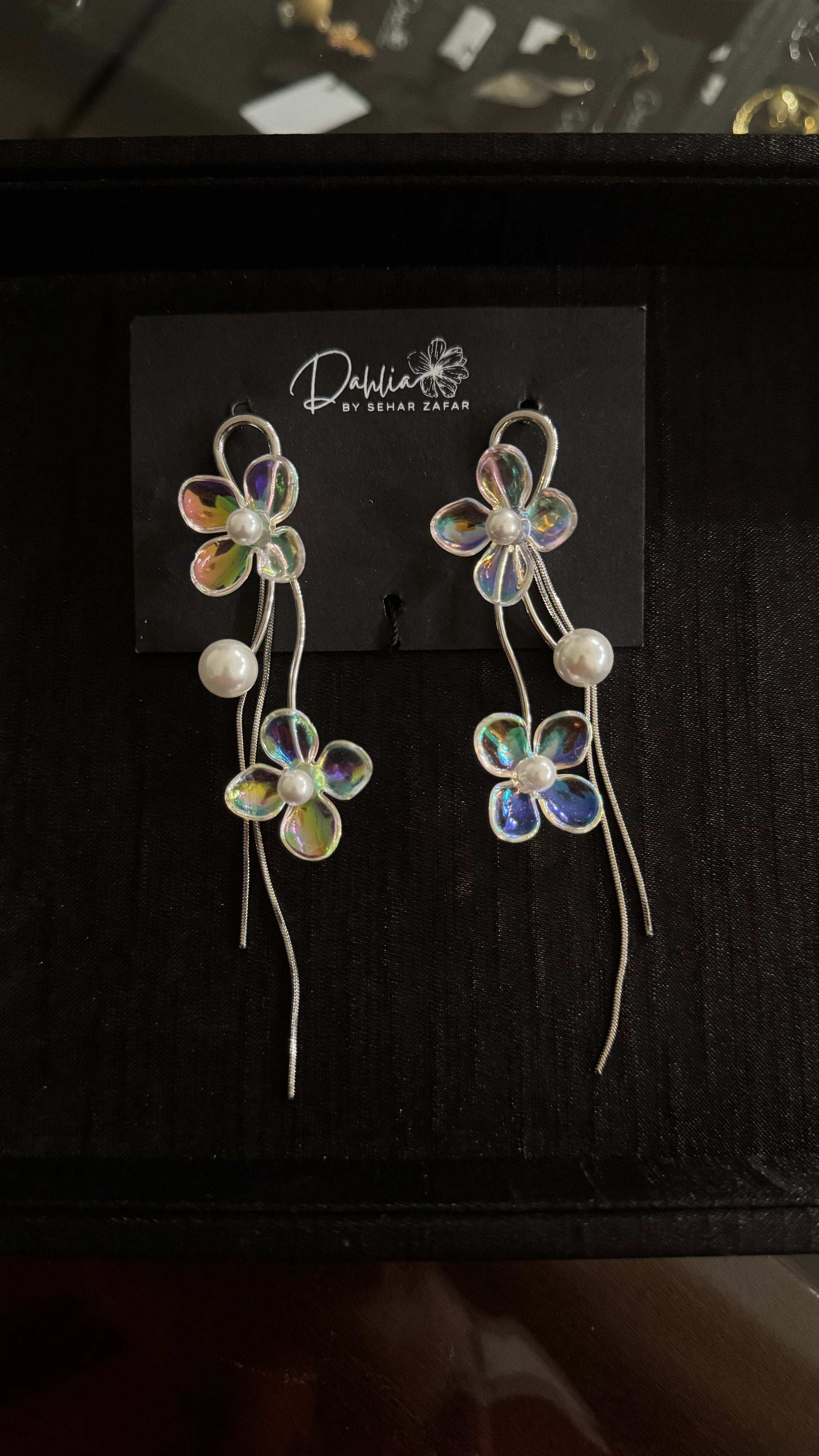 Iridescent floral earrings with pearl accents and curved metallic strands on black branded card