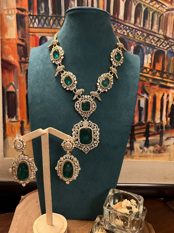 24 karat gold Kundan necklace with large emerald stones and matching earrings displayed on a teal stand.