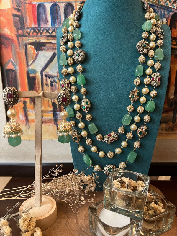 A luxurious gold-plated necklace with multicolored polkis, zircon, emeralds, and rubies, displayed on a teal mannequin bust, accompanied by a wooden cross and dried flowers.