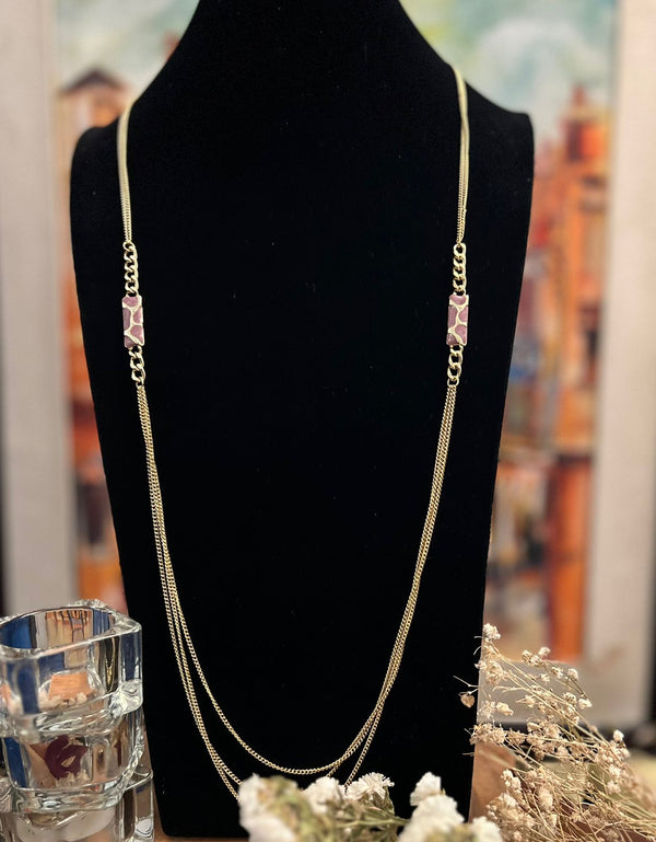 A gold-colored long necklace with a pink block-shaped pendant, displayed on a black mannequin against a background with floral decorations.