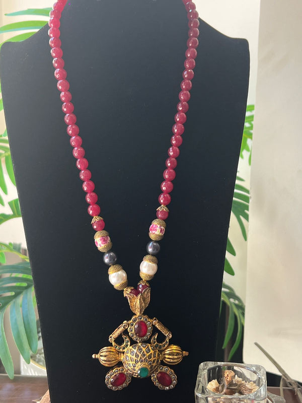 Long red beaded necklace with gold and gemstone pendant displayed on a black mannequin stand against a neutral indoor background