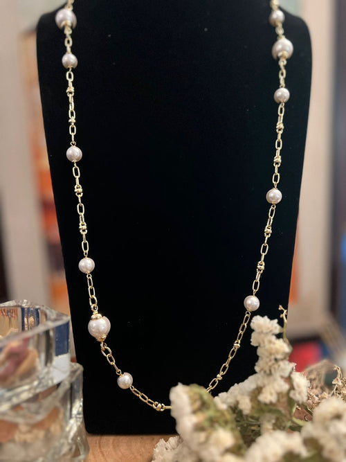 A gold-colored necklace with a chain design and pearl beads displayed on a mannequin bust, with a floral arrangement in the foreground.