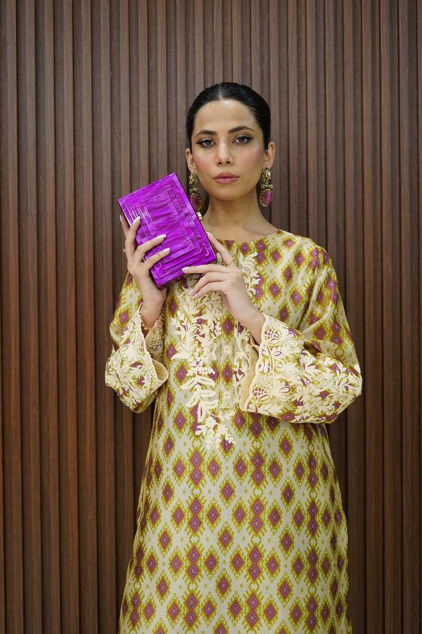 A person holding a purple square design clutch, with a patterned dress and earrings, standing against a wooden background.