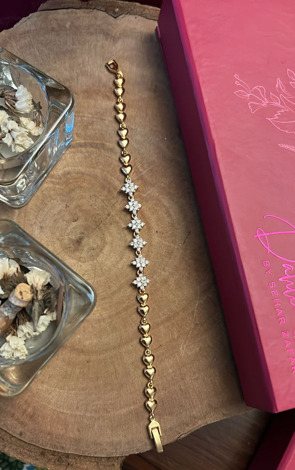 Gold heart chain bracelet with floral crystal center placed on a wooden surface beside a pink Dahlia by Sehar box