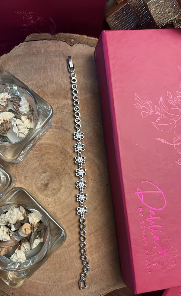 A sleek, silver bracelet with a series of connected metal links, displayed on a wooden surface next to a pink box with floral designs and a glass with mushrooms.