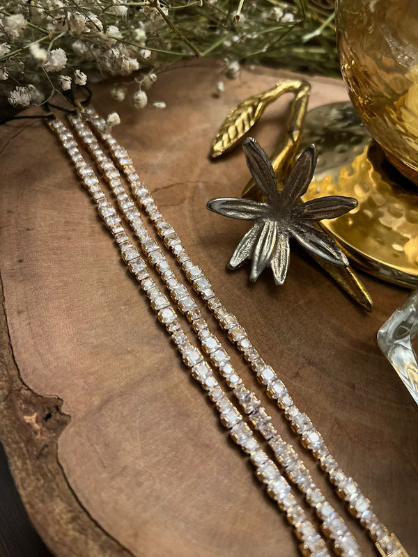 A gold tennis bracelet displayed on a wooden surface with decorative elements in the background.