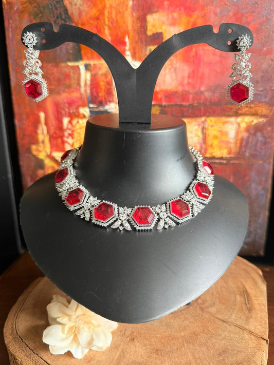 Jewelry set with red and silver necklace and earrings on a black mannequin against a warm-toned abstract background.