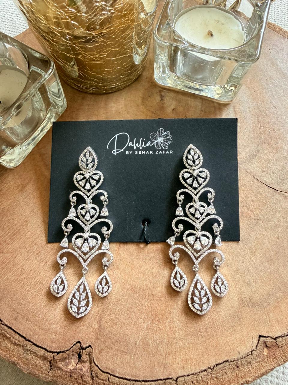 Silver earrings on a black card with 'Dahlia by Snehar Zafar' branding, placed on a wooden surface with candles.