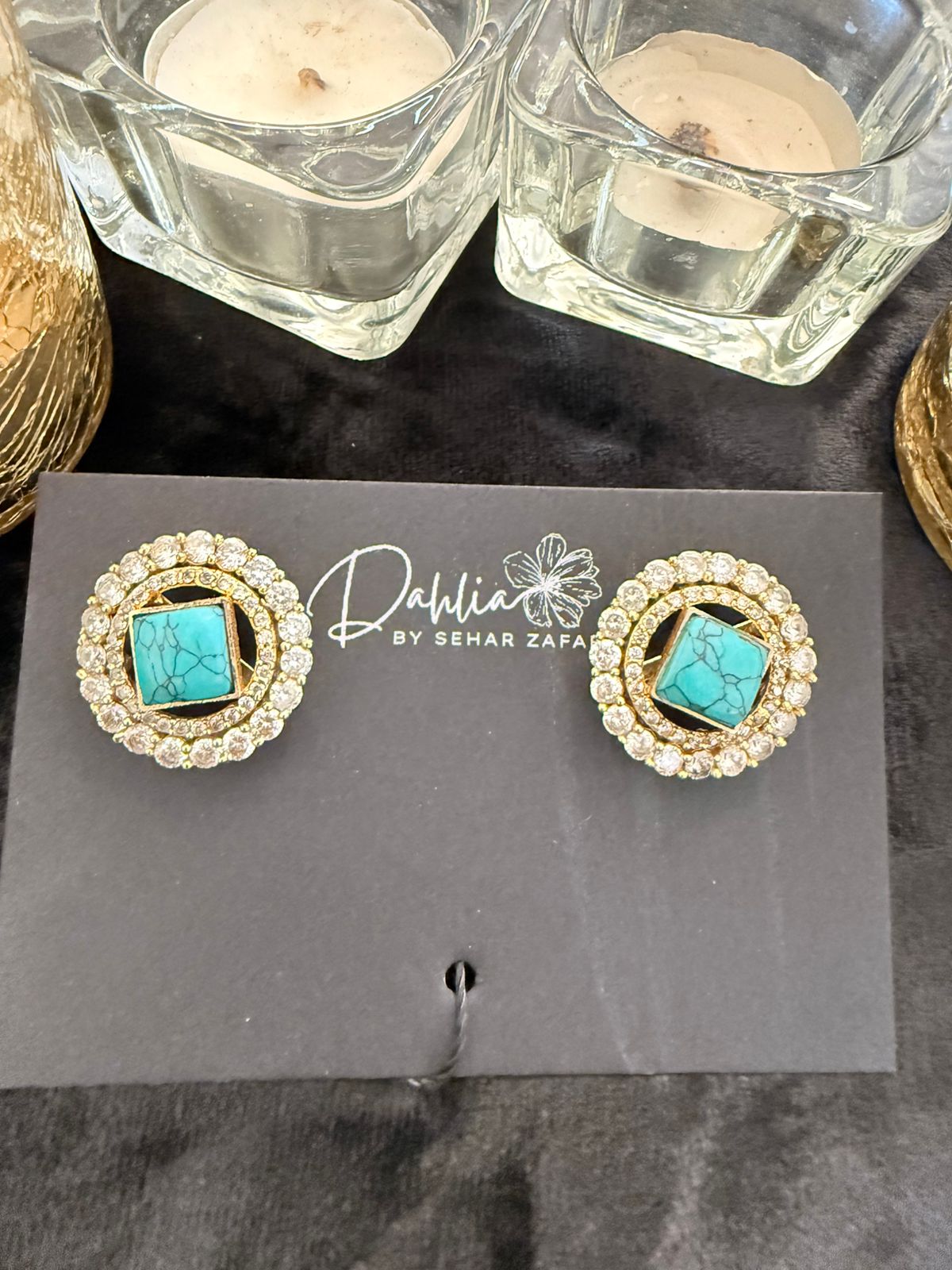 Pair of earrings with turquoise stones on a branded card, placed on a dark surface with candles in the background.