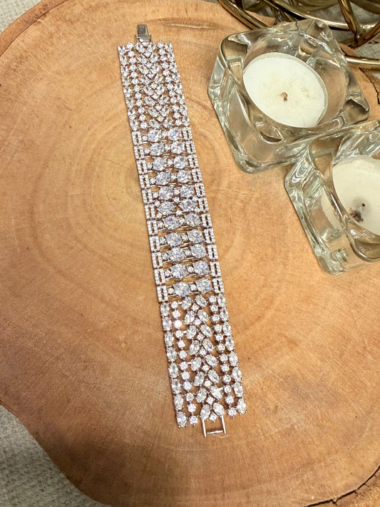 Silver bracelet with clear stones on a wooden surface next to a small candle in a glass holder.