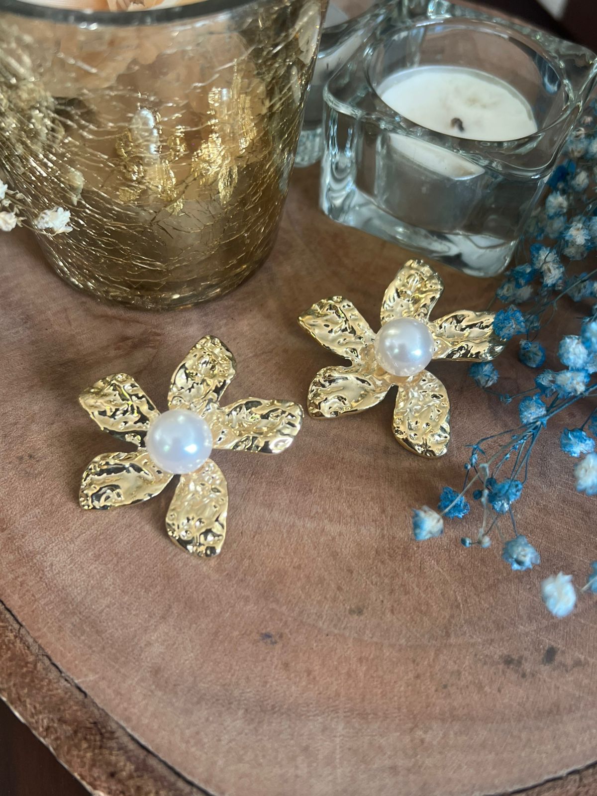 Gold flower-shaped earrings with pearls on a wooden surface with candles and flowers.