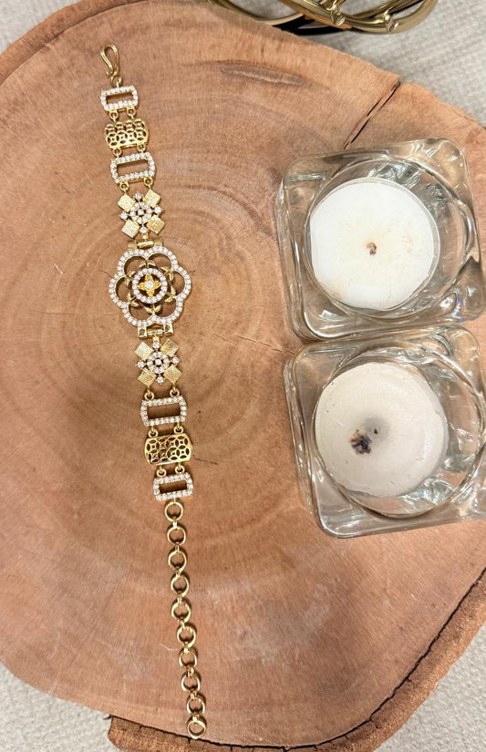 Gold bracelet with floral designs on a wooden surface with two small glass candle holders containing candles.