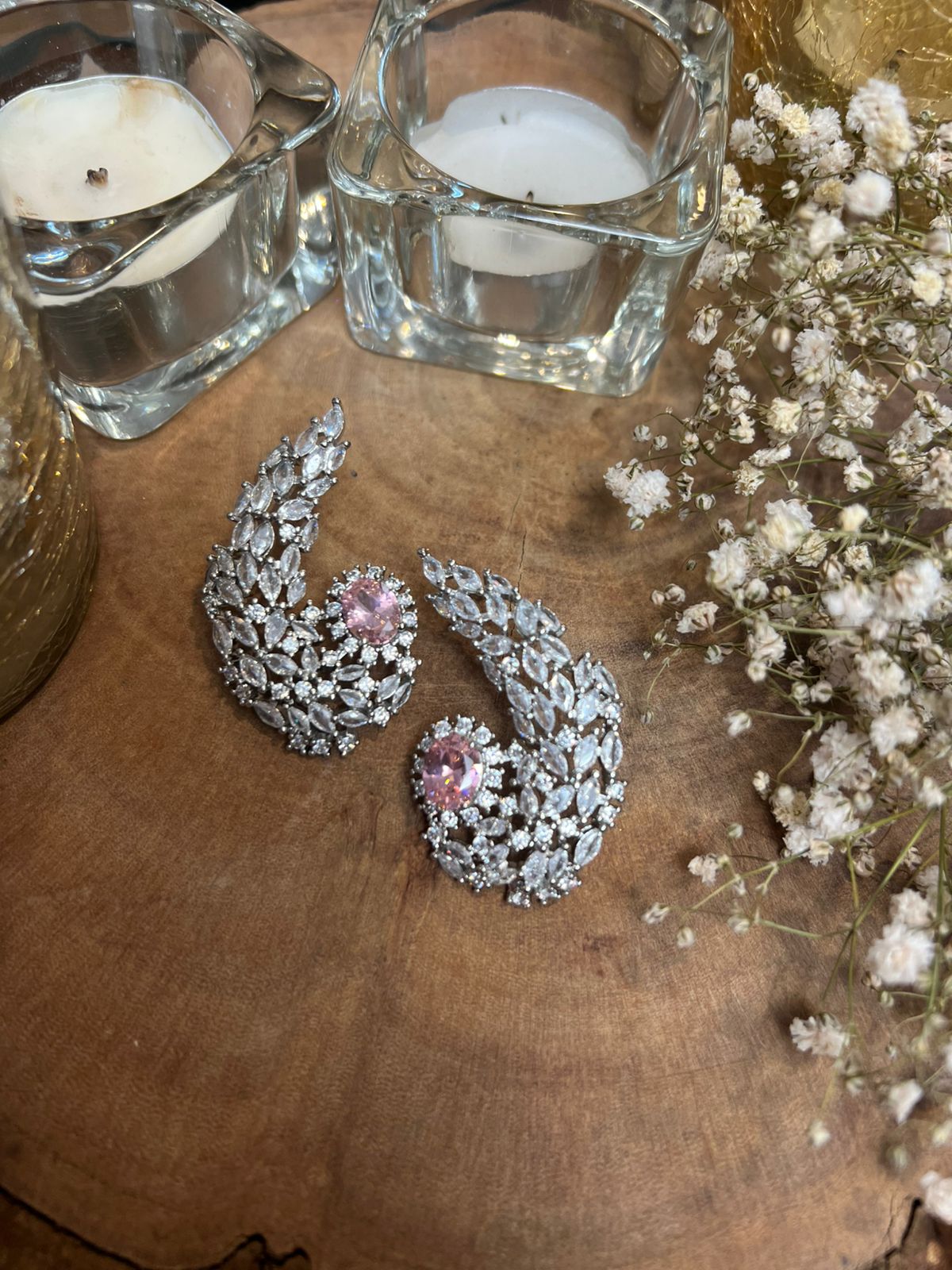 Pair of silver earrings with pink gemstones on a wooden surface with candles and flowers.