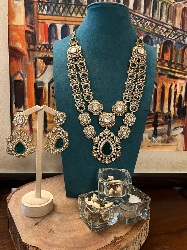 Double-layer Kundan necklace with emerald pendant and matching earrings displayed on a teal stand.