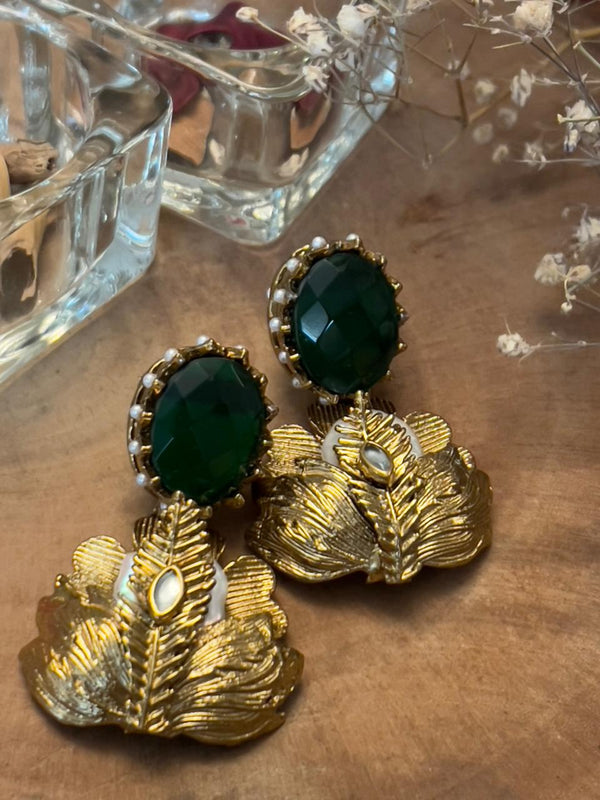 A pair of gold-toned earrings featuring green and white semi-precious stones, displayed alongside a glass dish with other jewelry and decorative items.